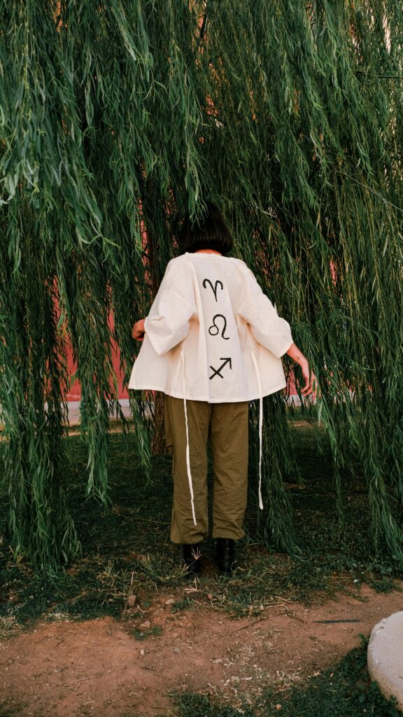 Woman wearing zodiac-themed coat standing under willow branches in a park.